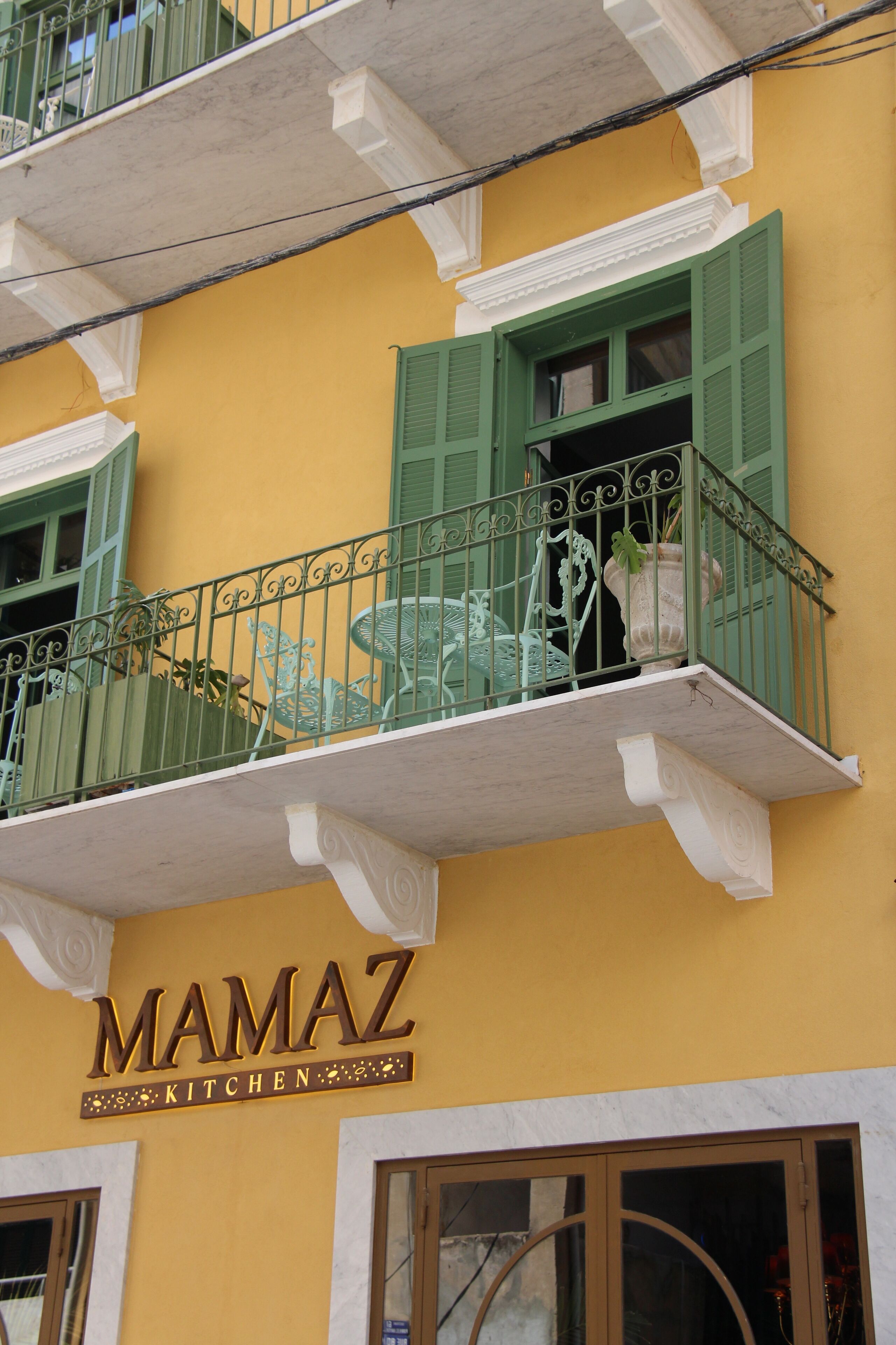 Un edificio con fachada amarilla, contraventanas verdes y un balcón con barandillas de metal. Se ve el letrero "MAMAZ KITCHEN".