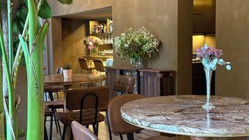 Un restaurante con mesas redondas y sillas de cuero marrón, una zona de bar con estantes llenos de botellas, y una planta verde grande.