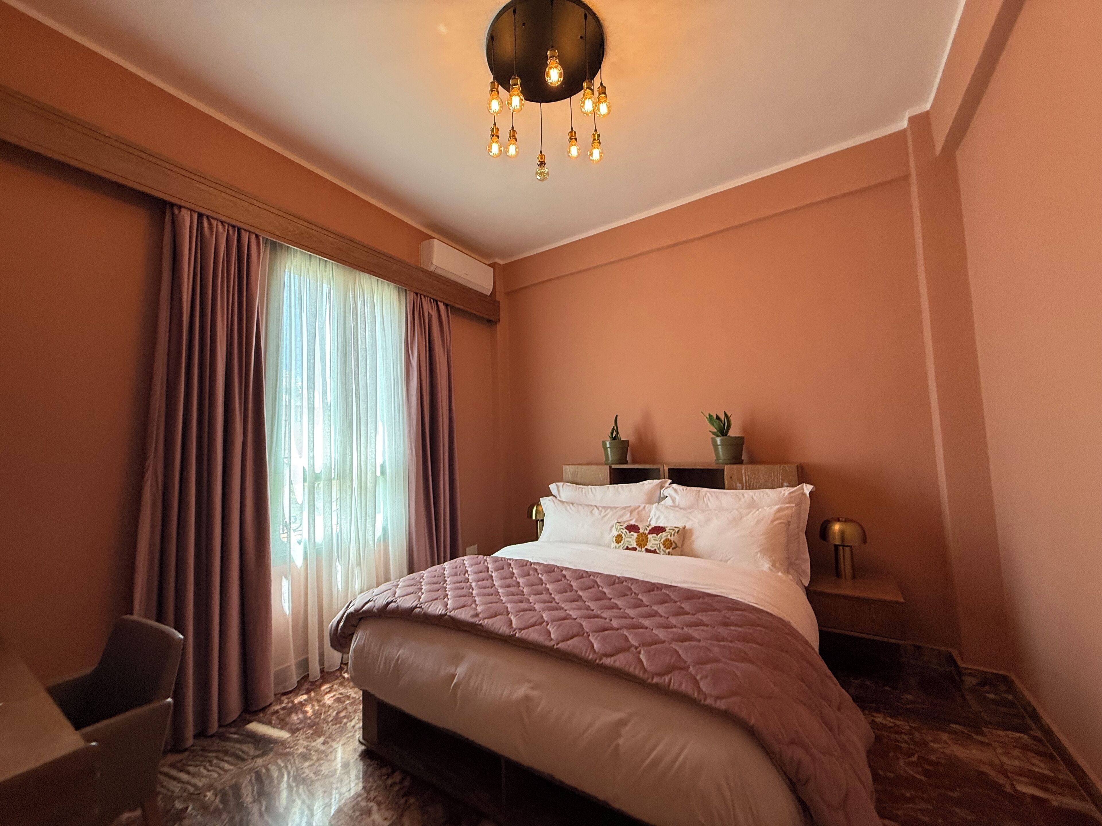 A hotel room with a large bed, a bedside table with a lamp, a potted plant, and a window with curtains.