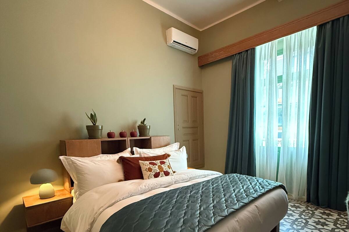 Une chambre à coucher avec un lit, des tables de chevet, une commode, une plante en pot, une fenêtre avec des rideaux et un sol à motifs.