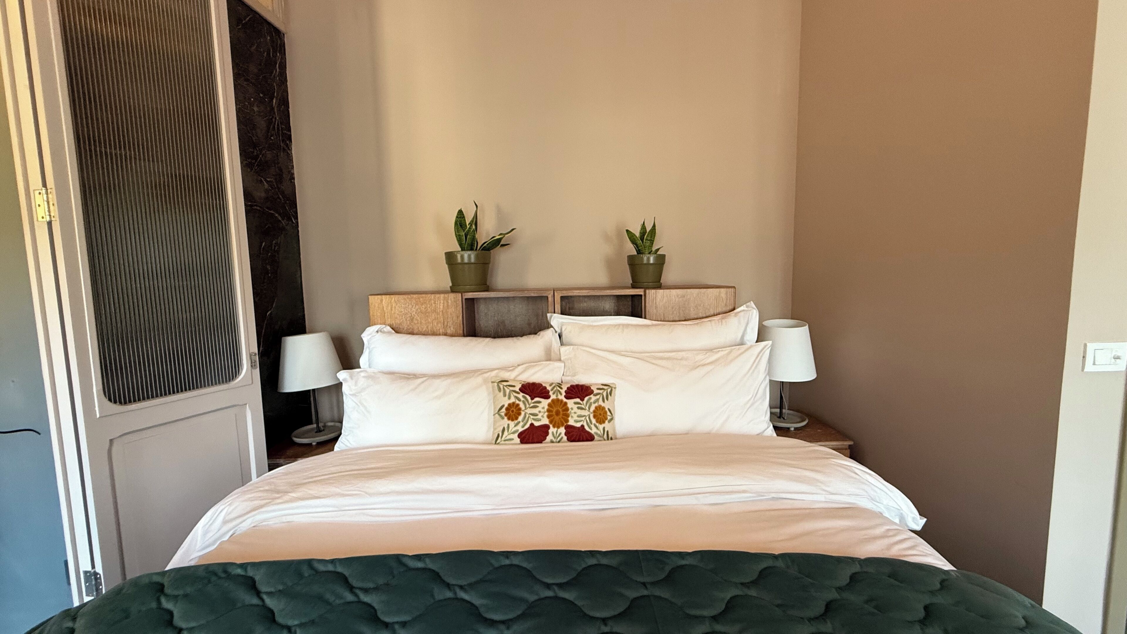A neatly made bed with a green upholstered headboard, two bedside tables with lamps, and two potted plants on the headboard.