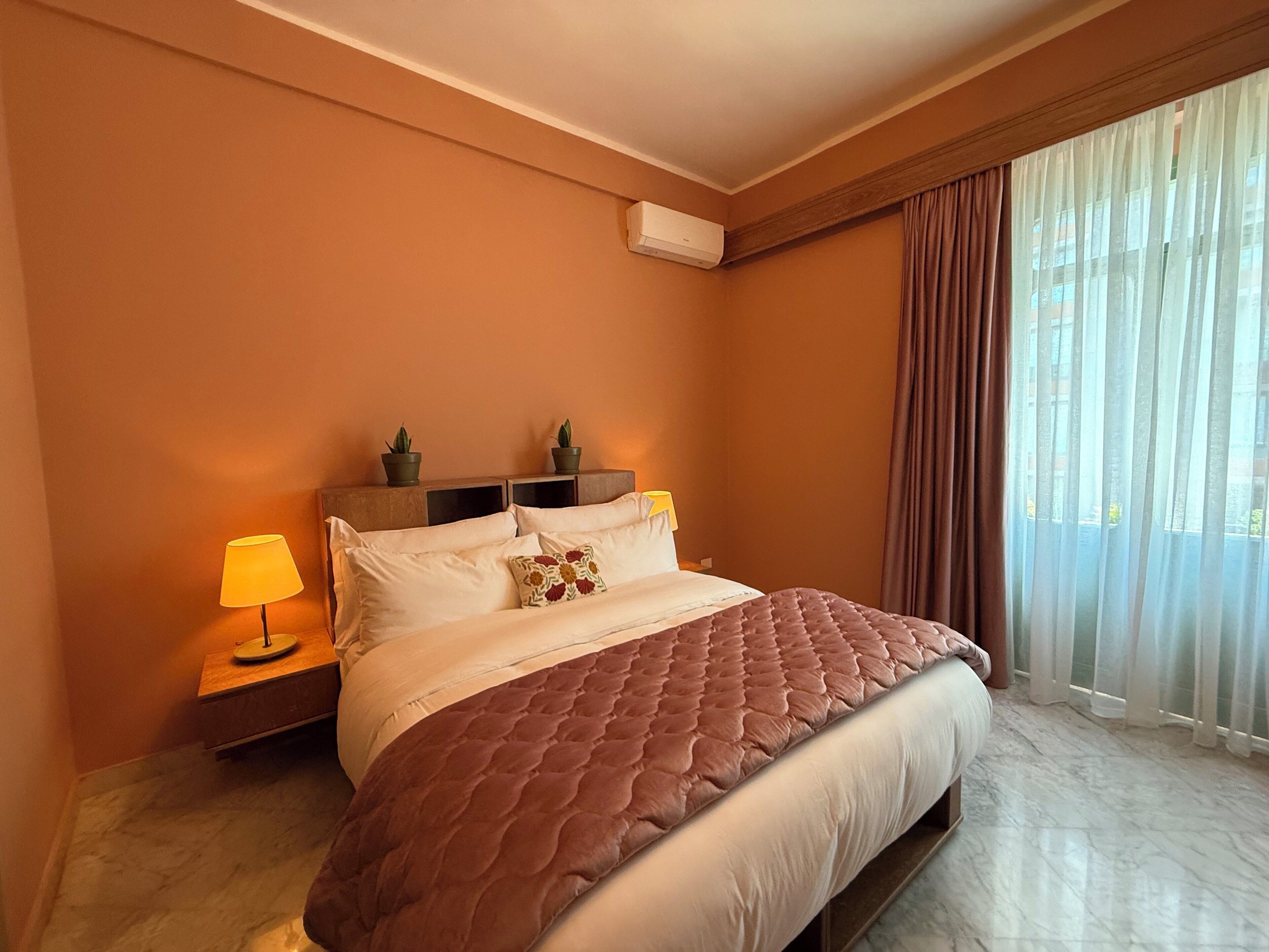 A hotel room with a large bed, two bedside tables with lamps, a potted plant, and a window with curtains.