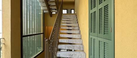 Una escalera de mármol con barandilla de metal, contraventanas verdes y una lámpara colgante.