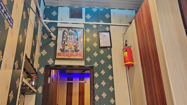 A room with patterned wallpaper, a ceiling light, a framed religious picture, a fire extinguisher, and a door leading to another room. - Ramlala Paying Guest House (Ayodhya)