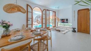 A modern dining area with a wooden table set for four, surrounded by wicker chairs. A large window offers a view of a swimming pool and outdoor seating.