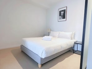 A minimalist bedroom with a single bed, white bedding, a small bedside table with a lamp, and a framed picture on the wall.