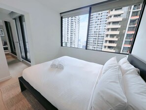 A modern hotel room with a large bed, white linens, a view of high-rise buildings, and a sliding door leading to a balcony.