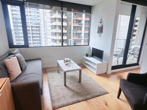 A modern living room with a sofa, a coffee table, a TV, and a large window offering a city view.