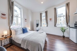 A modern hotel room with a large bed, a bedside table with a lamp, a wardrobe, a small kitchen area, and a potted plant. - Camberwell Studios, 10 mins to Station (London)