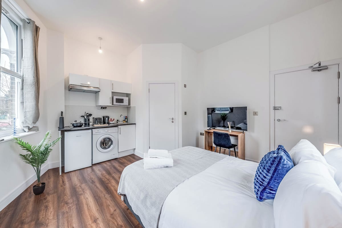 A modern hotel room with a bed, a desk, a TV, a washing machine, and a window with curtains.