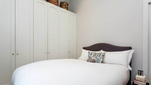 A bedroom with a large bed, white wardrobe, and a nightstand with books. - Gateway to Notting Hill, by Veeve (London)