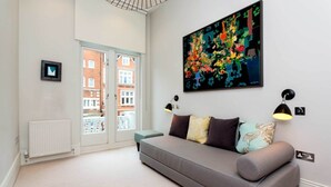 A modern living room with a grey sofa, colorful cushions, a large abstract painting, and a view through a glass door to a brick building. - Gateway to Notting Hill, by Veeve (London)