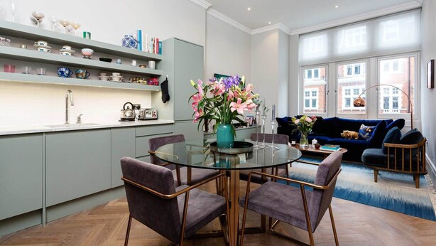 A modern kitchen with grey cabinets, a round glass table, and purple chairs. There is a dining area with a blue sofa and a dog. - Gateway to Notting Hill, by Veeve (London)
