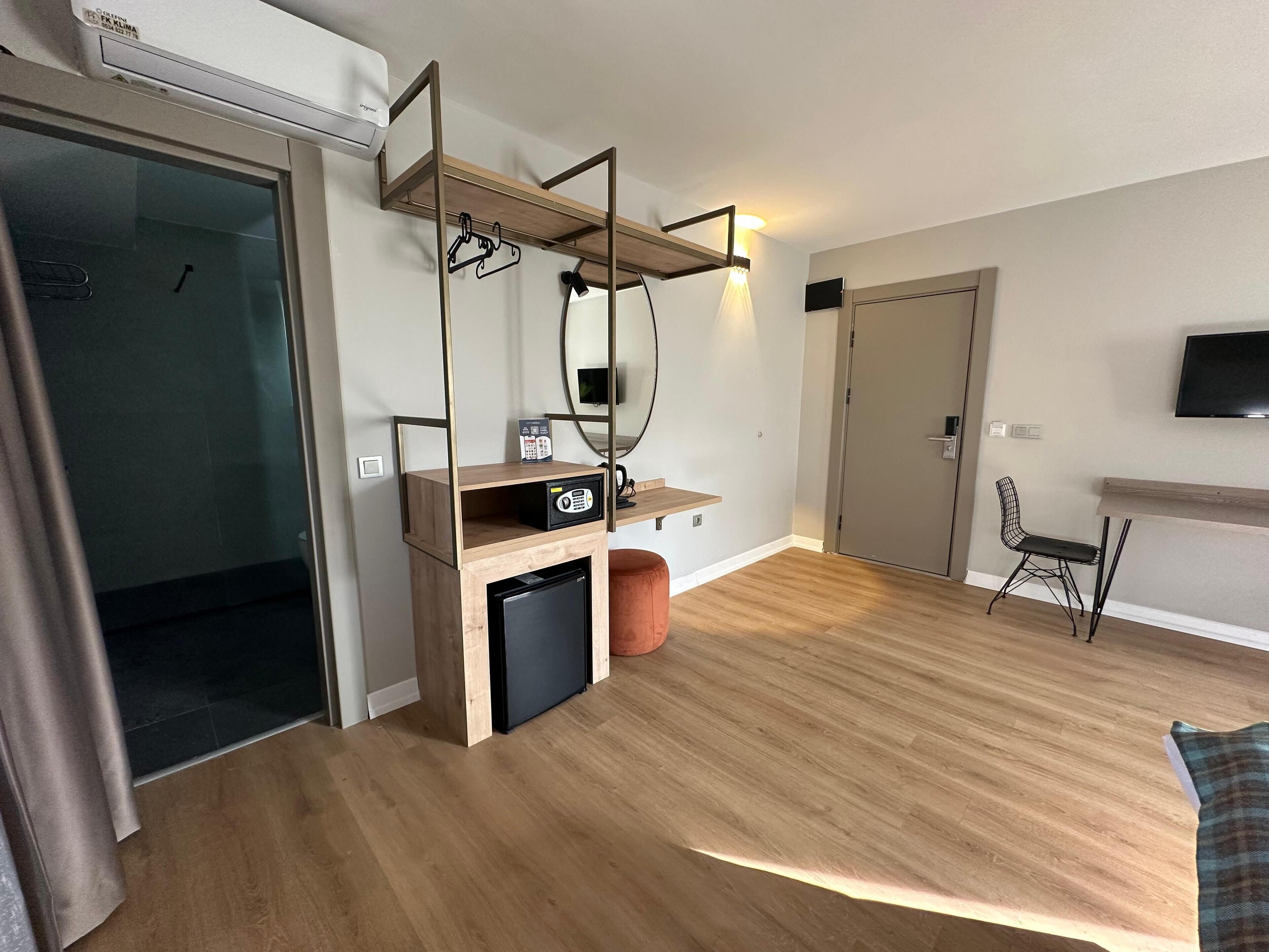 A modern hotel room with a wooden floor, a built-in desk with a chair, a small fridge, a mirror, and a TV mounted on the wall.
