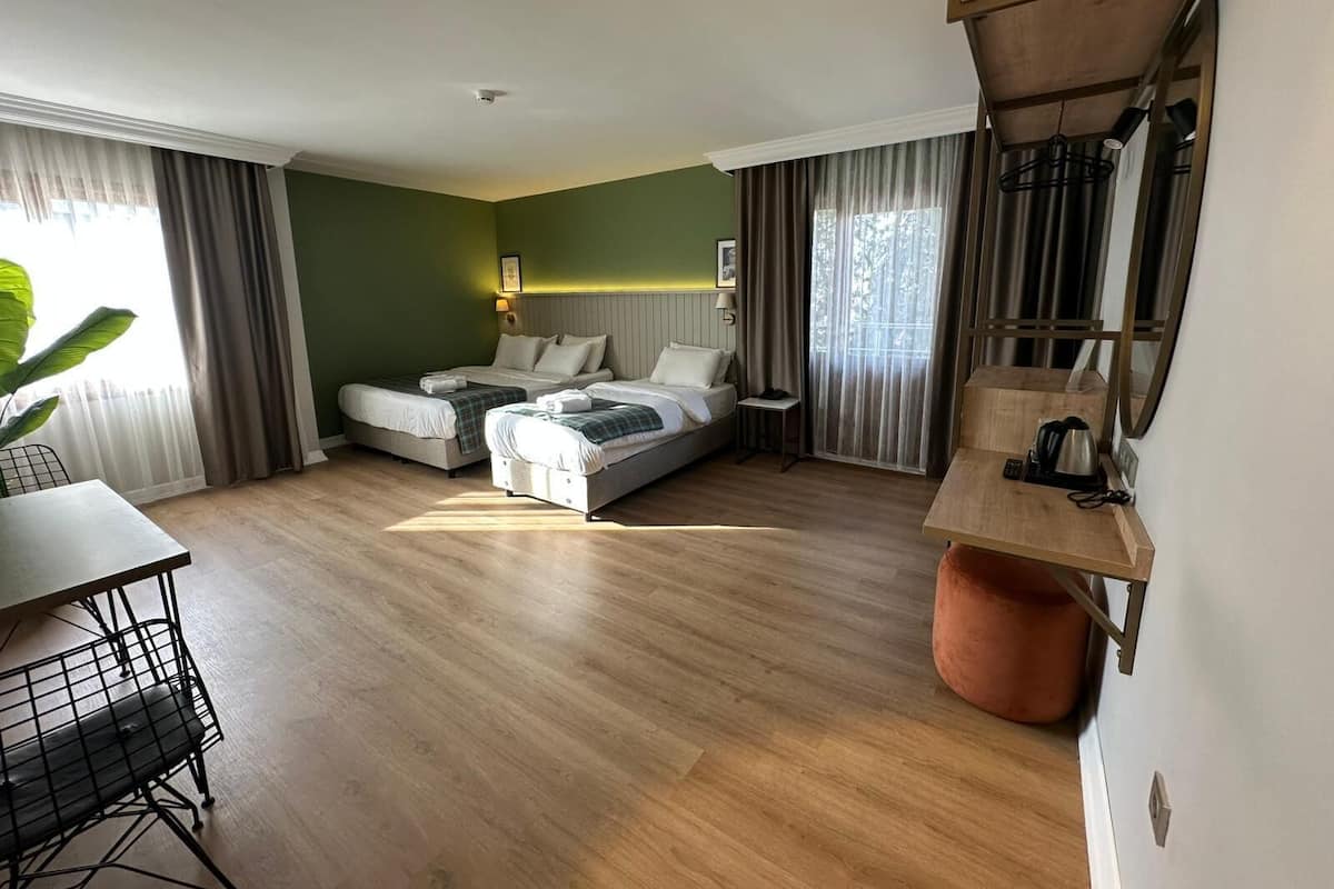 Habitació d'hotel amb dos llits, una taula de menjador, una cadira, una taula petita amb una tetera i una finestra gran amb cortines.