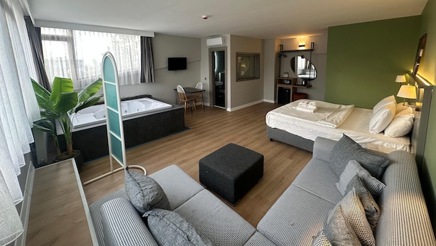 Ein modernes Hotelzimmer mit einem großen Bett, einer Couch, einem kleinen Tisch, einem Schreibtisch, einem Fernseher, einem Spiegel, einem Badezimmer und einem großen Fenster.
