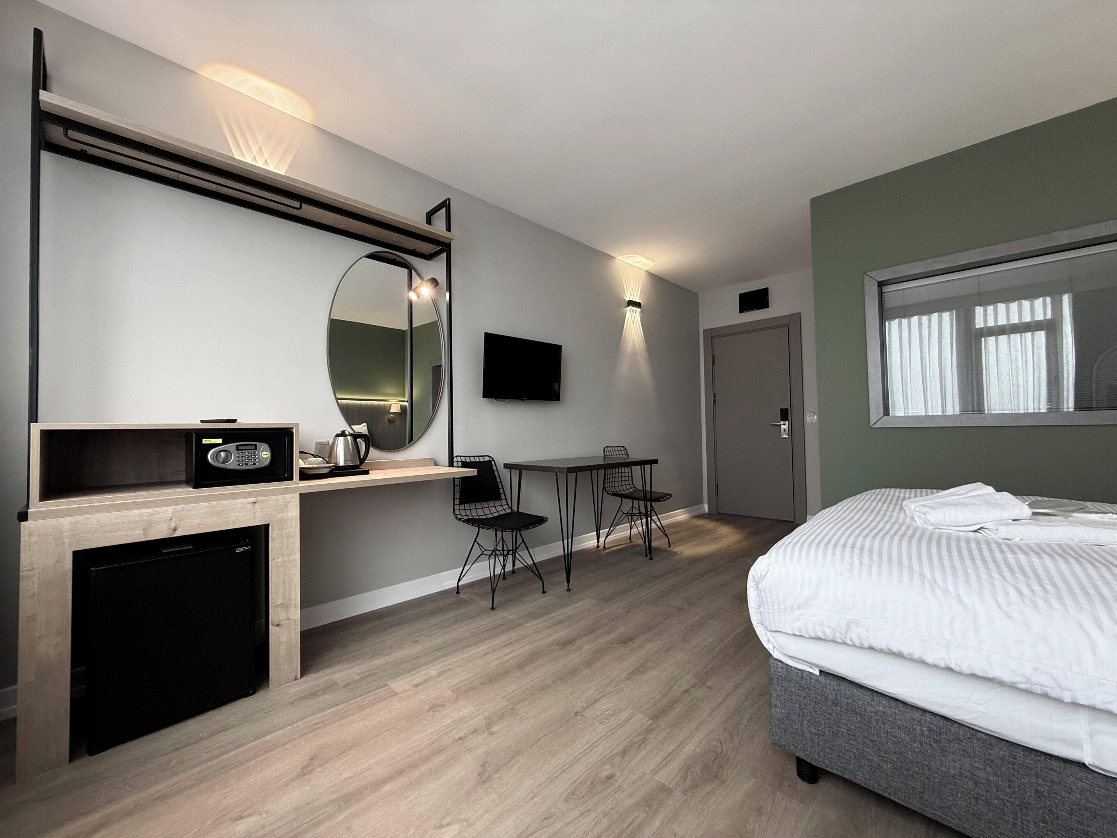 Habitació d'hotel moderna amb un llit, una taula de treball, una cadira, un televisor, un mirall i una cuineta petita.