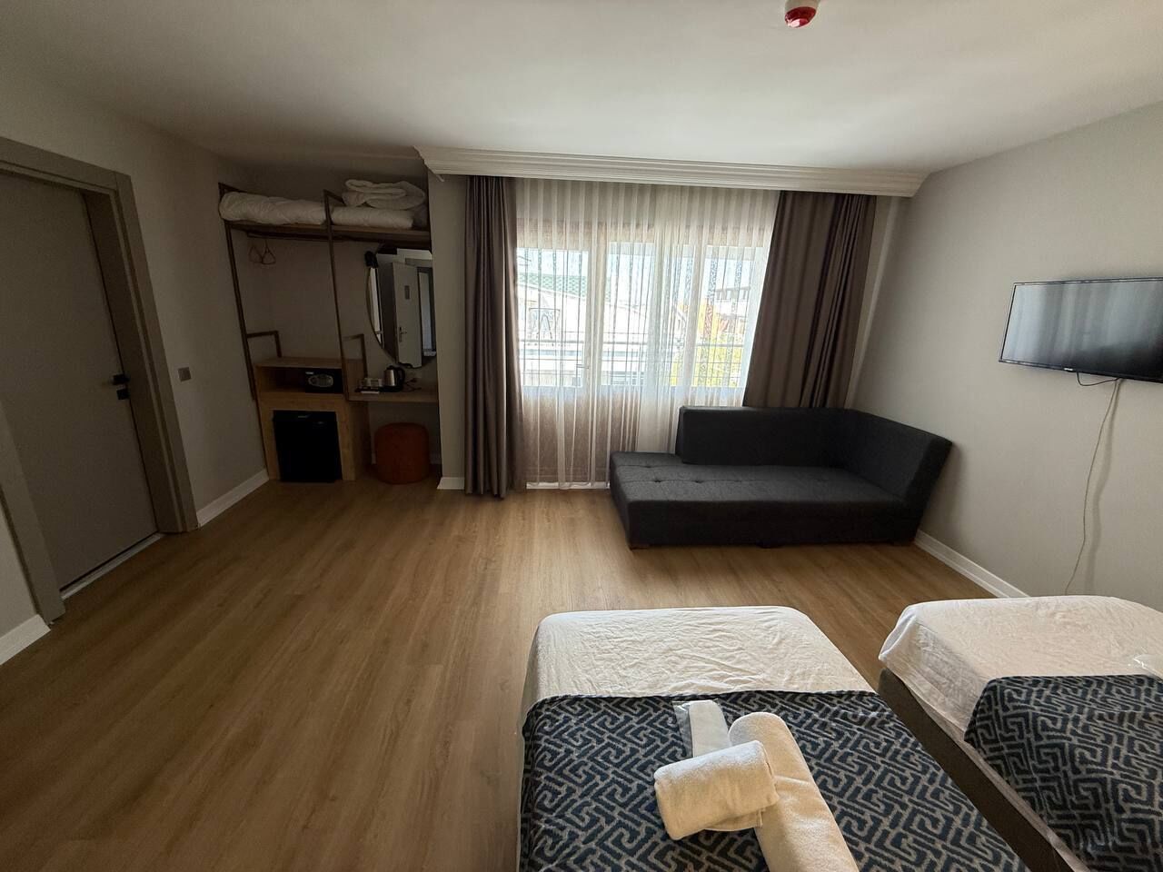 Une chambre d’hôtel avec deux lits simples, un canapé, une télévision, un bureau avec un ordinateur et une fenêtre avec des rideaux.