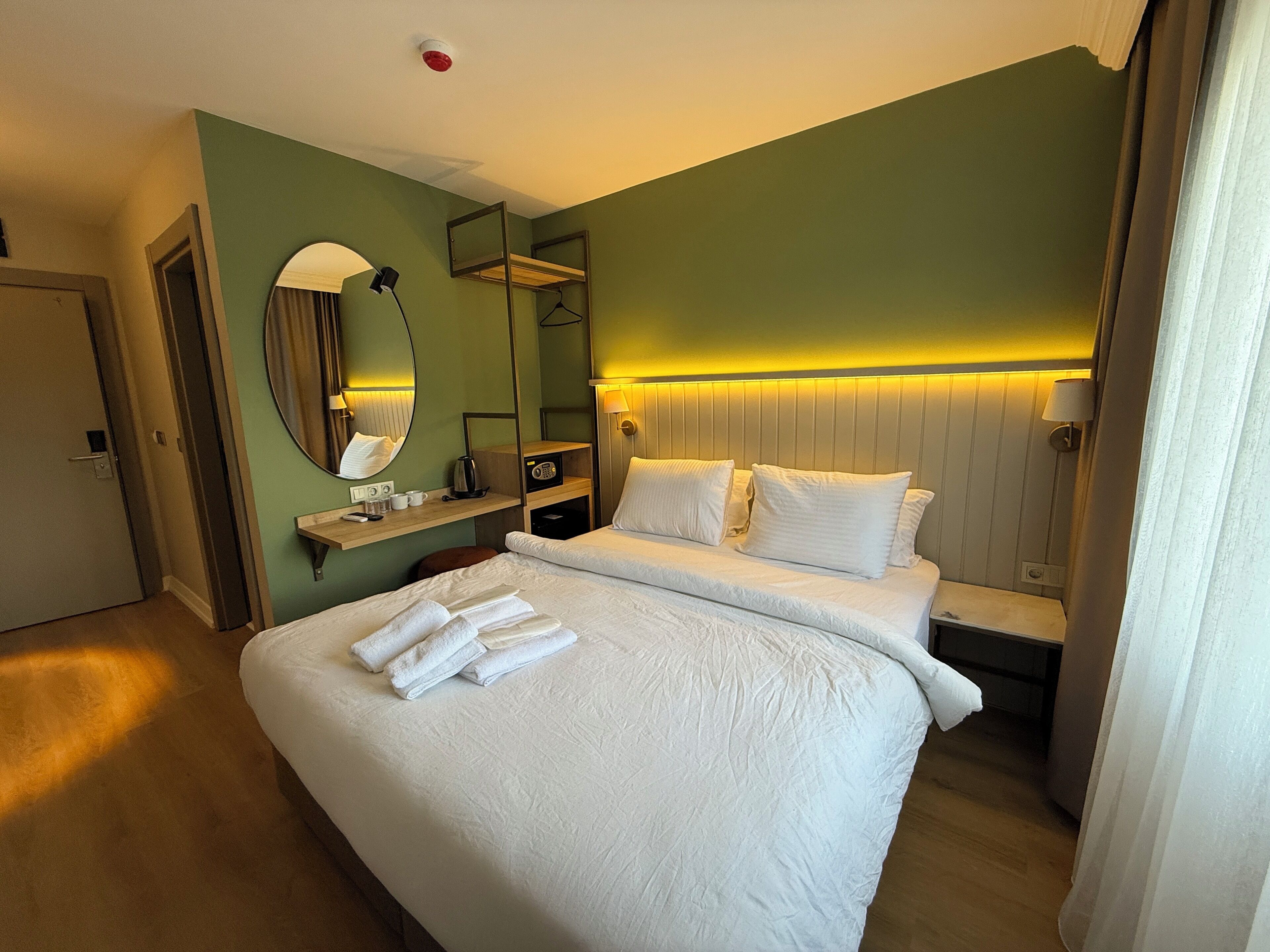 A hotel room with a large bed, a bedside table, a mirror, a lamp, and a wall-mounted shelf.