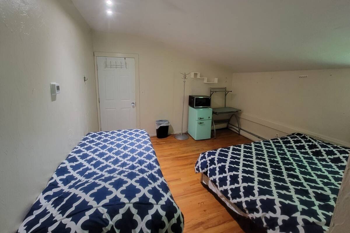 Quarto pequeno e limpo, com duas camas, uma escrivaninha, uma cadeira e um cesto de lixo.