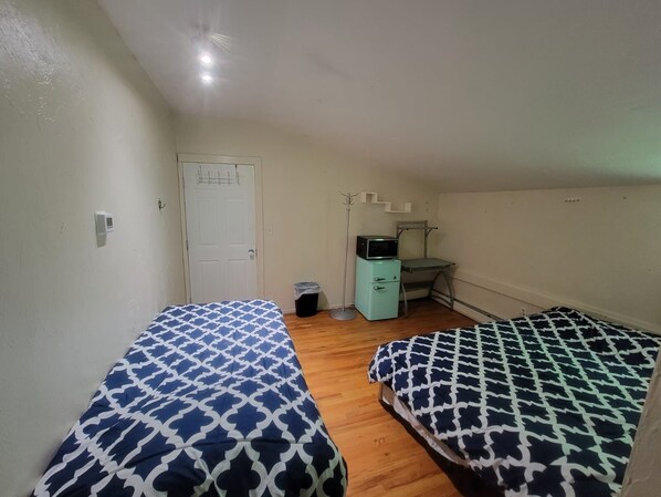 A small, clean room with two beds, a desk, a chair, and a trash bin.