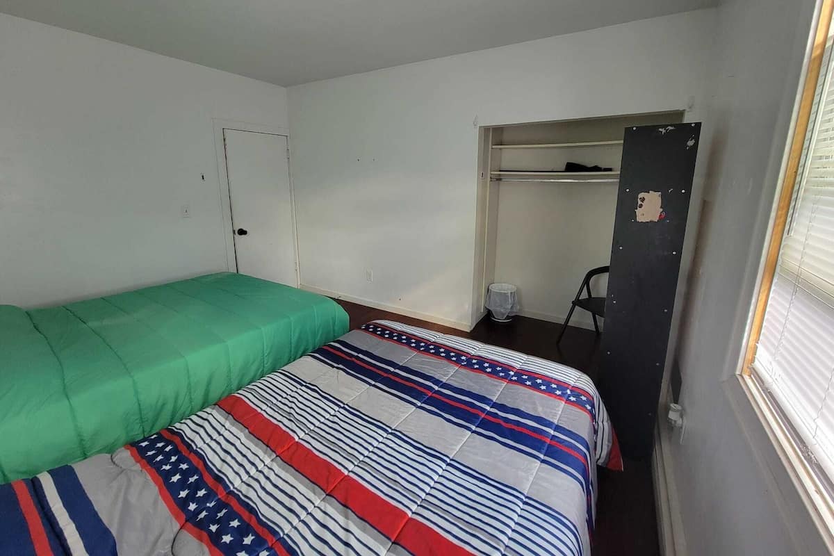 Quarto pequeno com uma cama de solteiro, colcha verde e manta listrada. Há um guarda-roupa com a porta entreaberta, uma cadeira e uma mesa pequena.