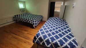 A small, single-room apartment with two beds, wooden flooring, a window with blinds, and a visible electrical outlet.