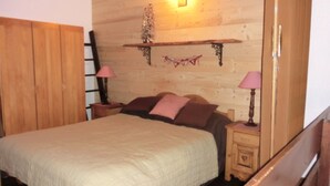 A bedroom with a wooden headboard, a bed with a quilt, two bedside tables with lamps, and a wall-mounted shelf with a decorative item. - Duplex 6 Guests, Near Ski Slopes, Balcony, Pet-Friendly (Saint-Chaffrey)