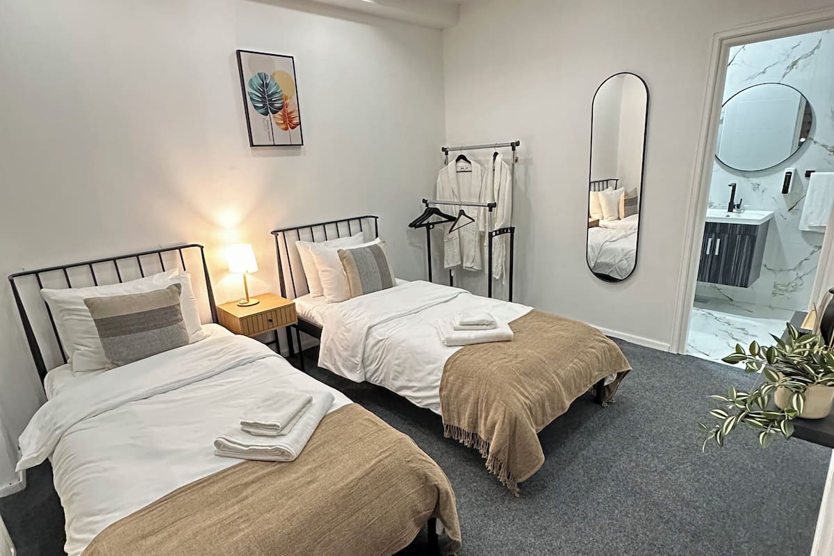 Une chambre d’hôtel avec deux lits, une table de chevet avec une lampe, un porte-vêtements, un miroir et une vue sur la salle de bain.
