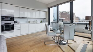 Una cocina moderna con armarios blancos, horno empotrado y un comedor con mesa de vidrio y sillas de metal.