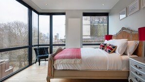 Un dormitorio con un ventanal grande, una cama con ropa de cama blanca, un sillón oscuro y una mesita de noche con una lámpara.