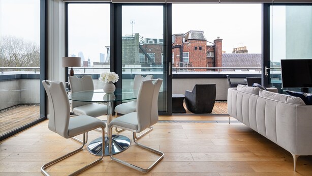 Una sala de estar moderna con una mesa de vidrio, sillas blancas y un sofá. Ventanas amplias ofrecen vistas del paisaje urbano.
