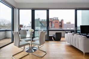 Una sala de estar moderna con una mesa de vidrio, sillas blancas y un sofá. Ventanas amplias ofrecen vistas del paisaje urbano.