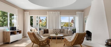 A modern living room with a sofa, two wicker chairs, a coffee table, a fireplace, and large windows with curtains.