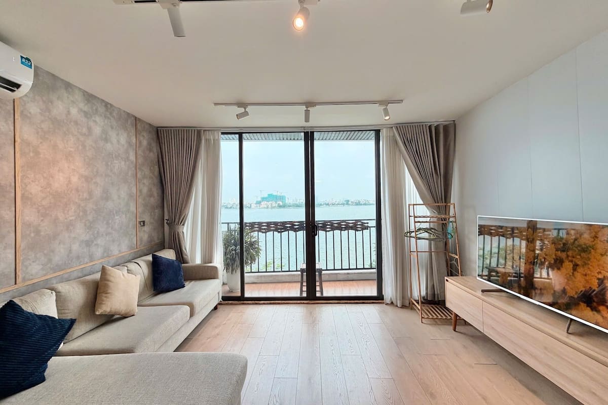 A modern living room with a sofa, a TV, and a view of a body of water through large glass doors.