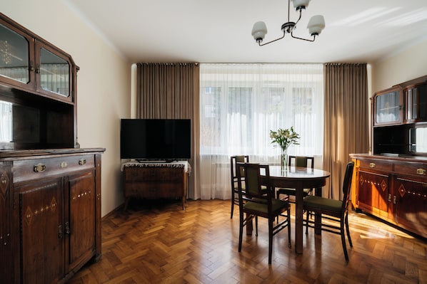 A room with a wooden herringbone floor, a dining table with chairs, a television, and a cabinet with a mirror.