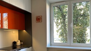 A modern kitchen with white cabinets, a red accent wall, a coffee maker, and a window with a view of trees. - Native Apartments Fałata 12 (Kraków)