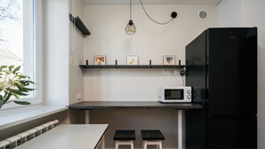 A compact kitchen with a black refrigerator, a white microwave, and a shelf with framed pictures. There is a window with a plant on the sill and a table with two stools. - Native Apartments Fałata 12 (Kraków)