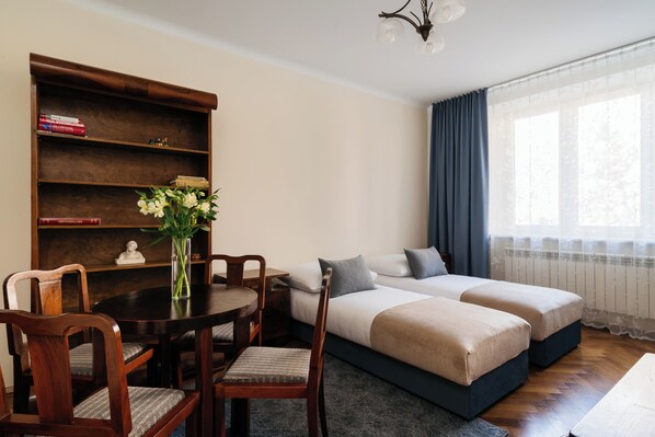A room with a wooden desk, chairs, a bookshelf, a vase with flowers, a bed with pillows, and a window with curtains. - Native Apartments Fałata 12 (Kraków)