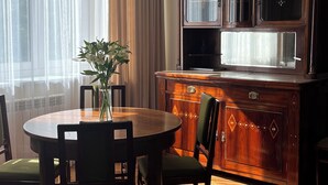 A dining area with a round wooden table, green upholstered chairs, a vase with white flowers, and a wooden cabinet with glass doors. - Native Apartments Fałata 12 (Kraków)