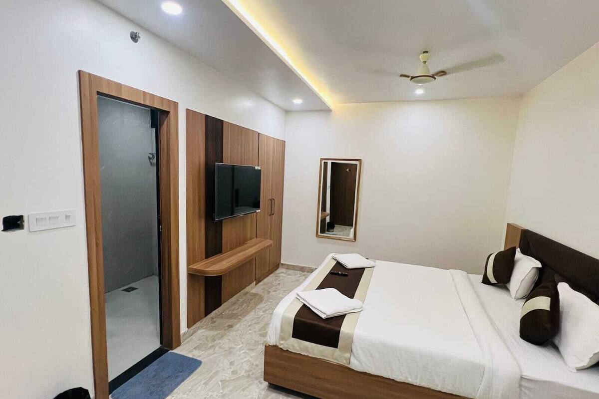 Un dormitorio con cama, televisor montado en un mueble de madera, ventilador de techo y un espejo pequeño en la pared.
