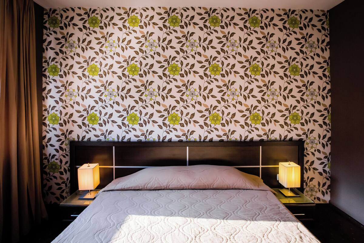Una cama bien hecha con un estampado floral en el papel tapiz, dos lámparas de noche y un cabecero oscuro.