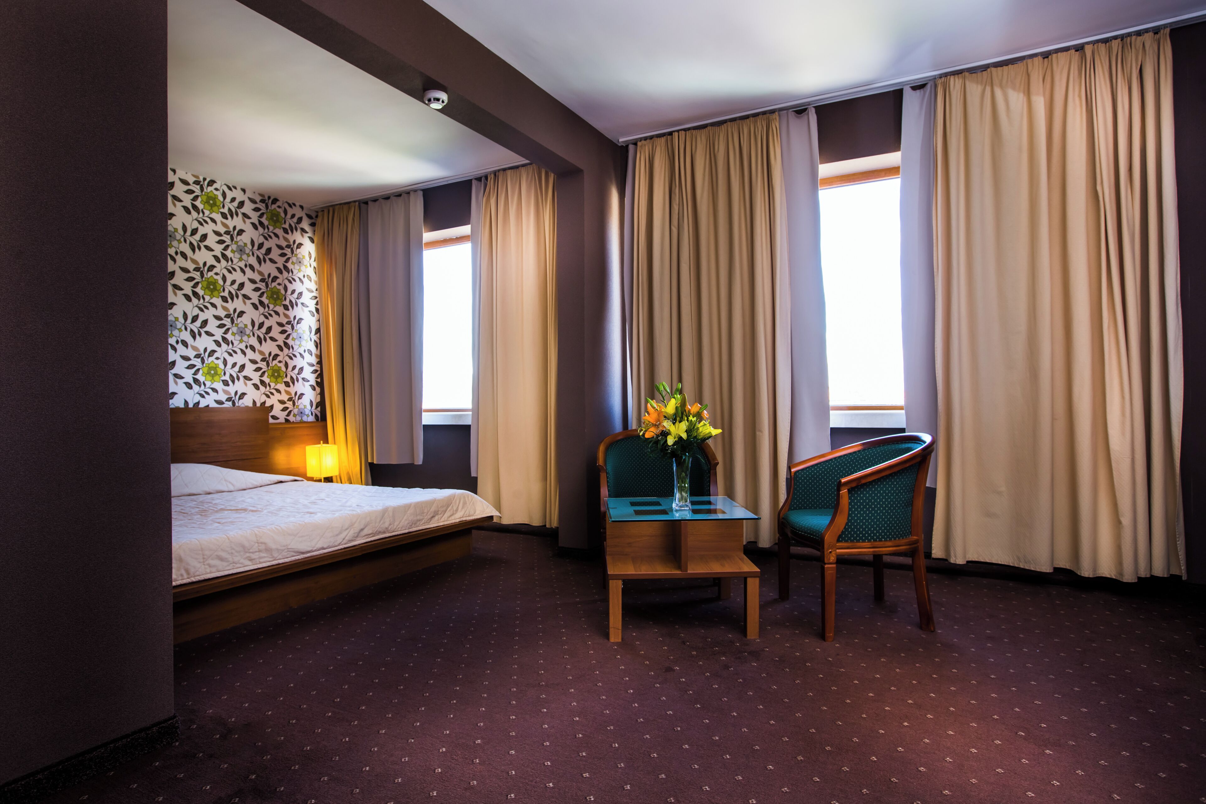 A hotel room with a bed, a desk with a vase of flowers, a chair, and curtains.