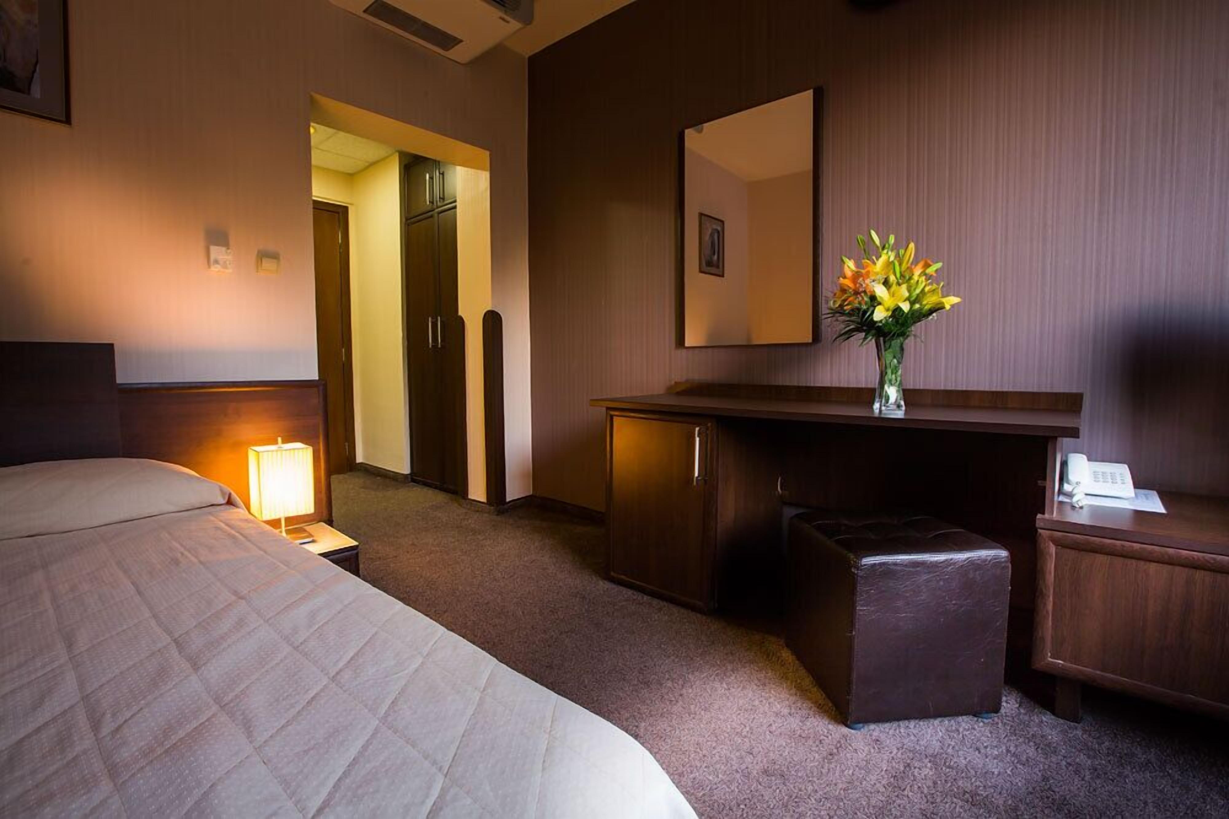 Habitación de hotel con cama, mesita de noche con lámpara, escritorio con espejo, armario pequeño y un jarrón con flores.