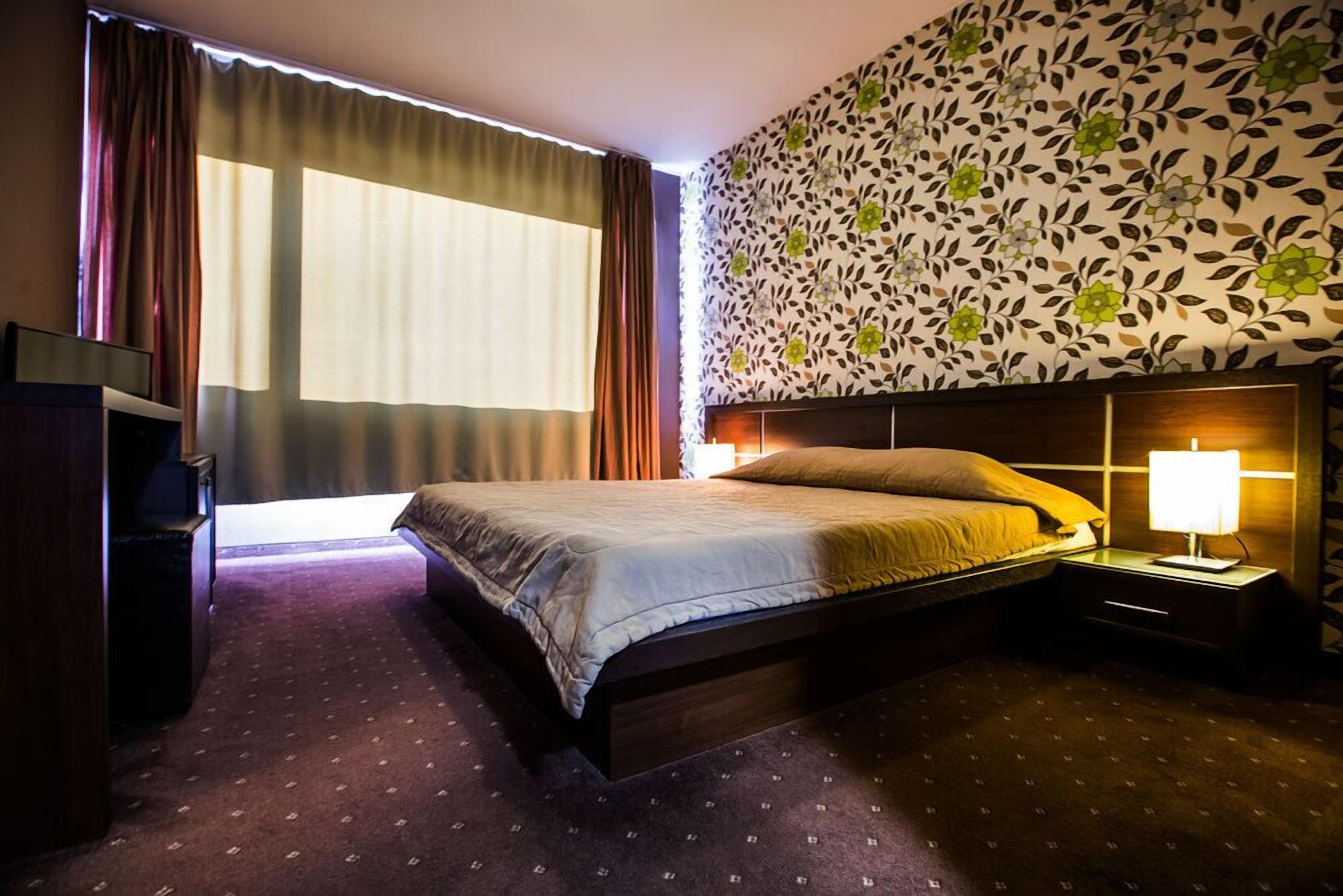 A hotel room with a large bed, a nightstand with a lamp, a wardrobe, and a window with curtains.