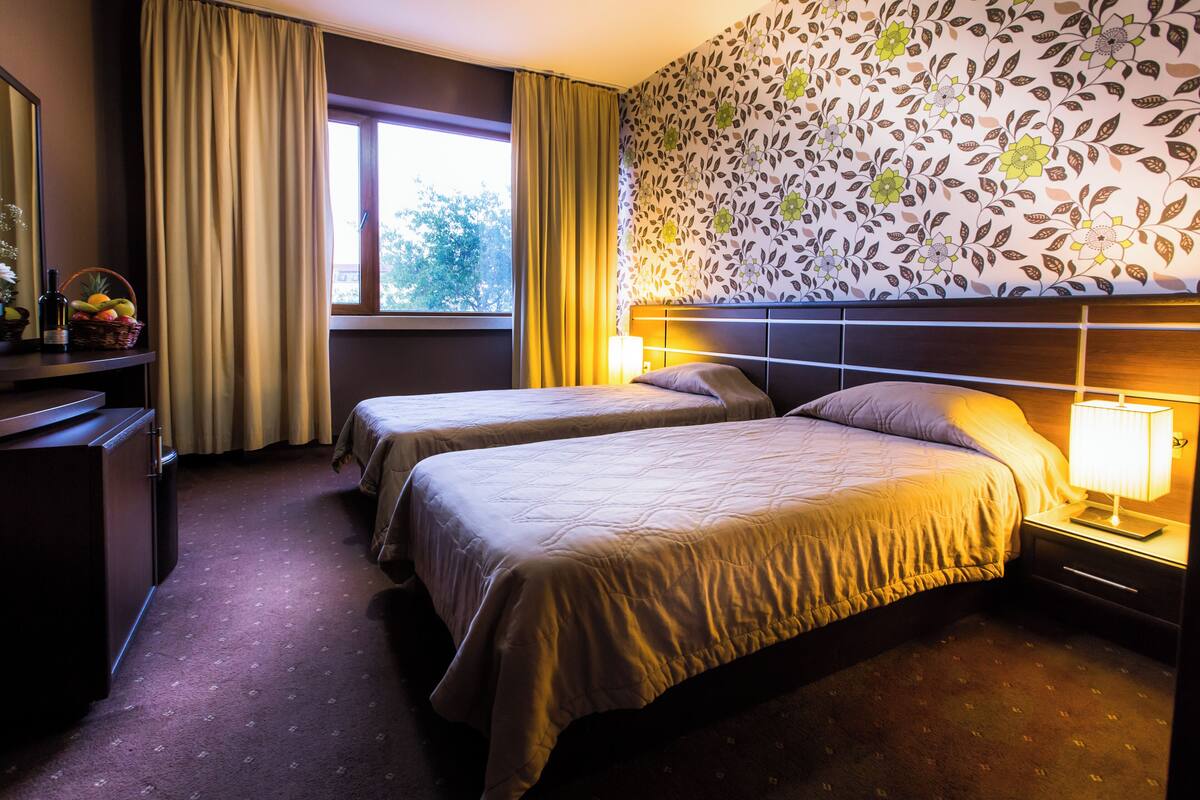 Habitación de hotel con dos camas, una mesita de noche con lámpara, cómoda y ventana con cortinas, además de un papel tapiz floral.