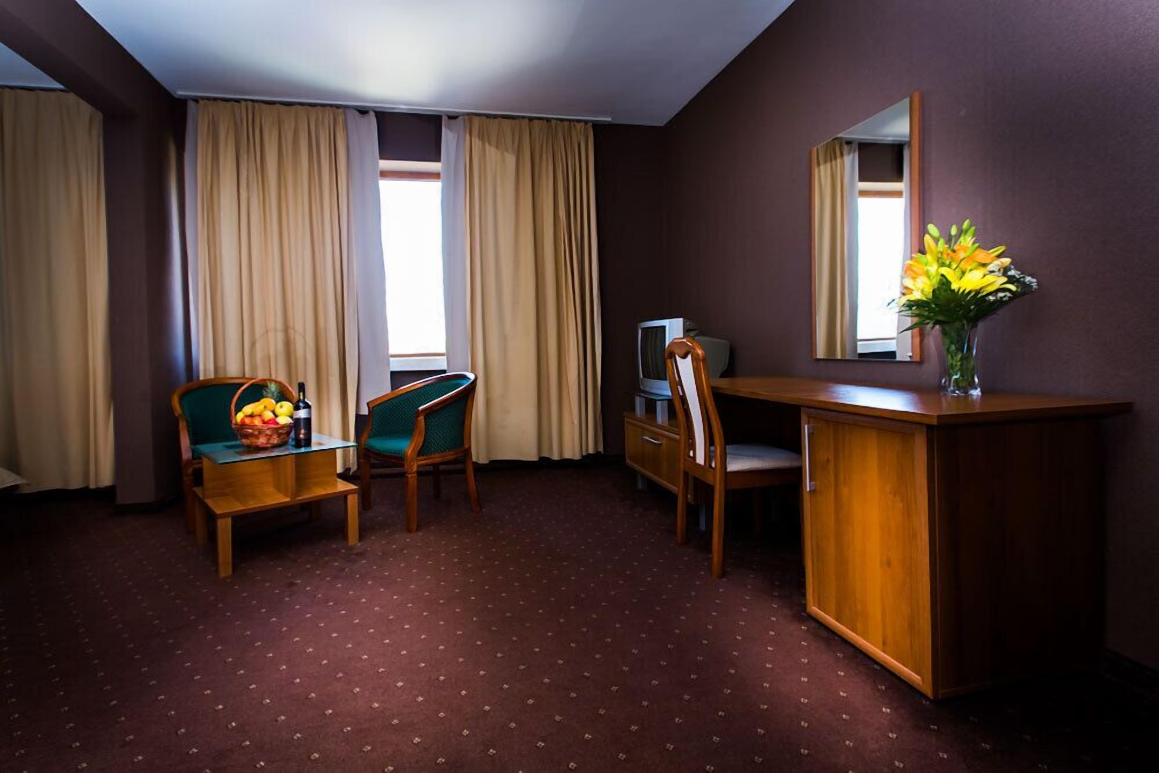 Une chambre d’hôtel avec un bureau en bois, deux chaises, une petite table sur laquelle se trouve un panier de fruits, une télévision et un vase avec des fleurs.