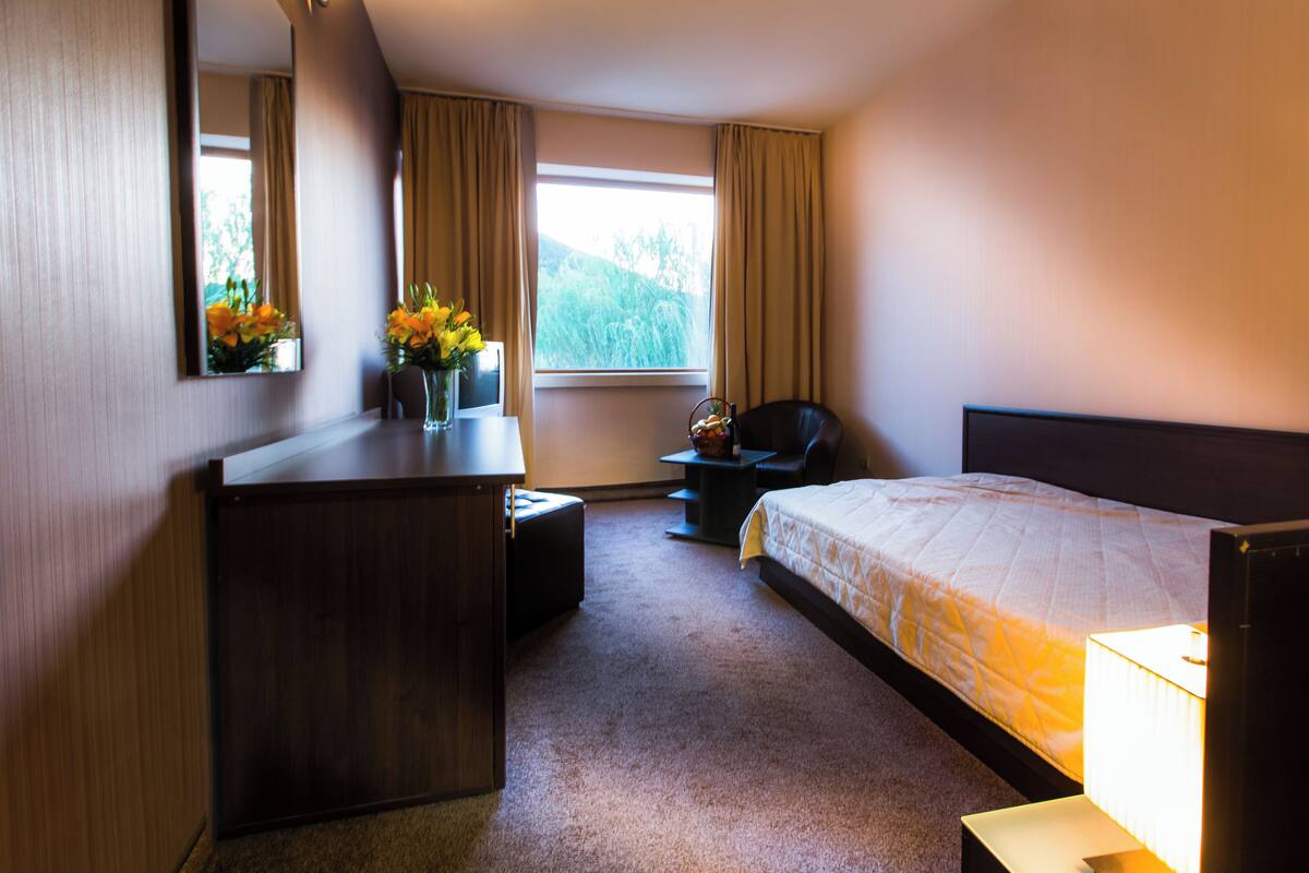 Una habitación de hotel con cama, escritorio, silla, lámpara y ventana con cortinas.