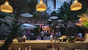 An outdoor bar area with hanging wicker lamps, umbrellas, and a stone wall backdrop. - Cali Stays & Cafe By the Bay (Vagator)