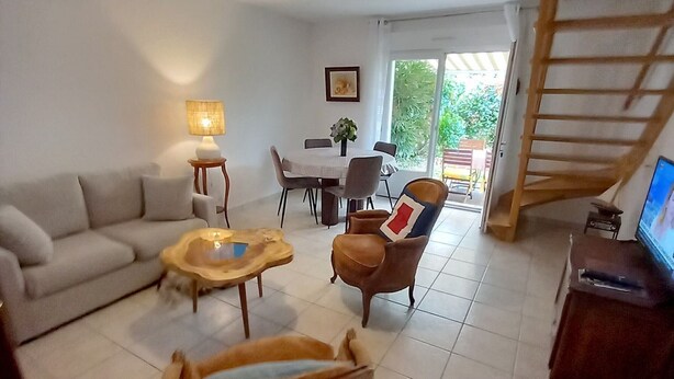 Sala de estar com sofá, mesa de centro e poltrona. Há uma área de jantar com mesa e cadeiras. Uma televisão está em um móvel. Uma escada é visível ao fundo.